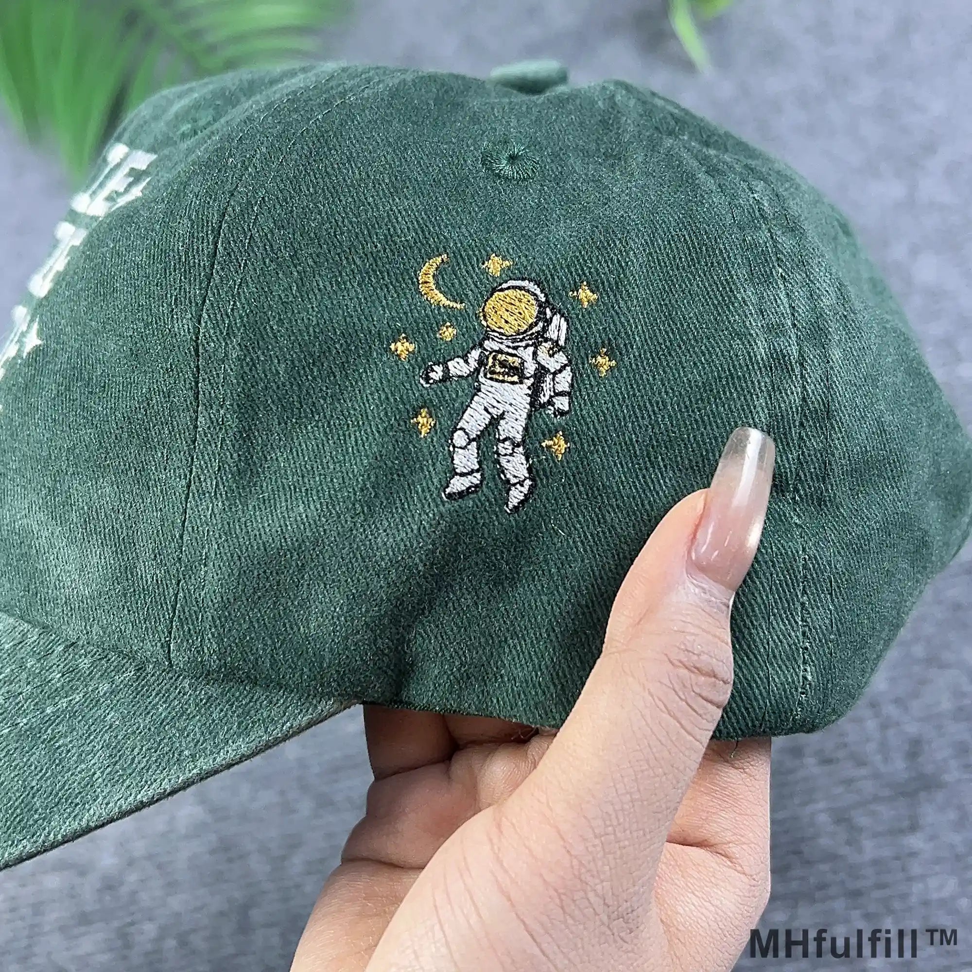 Detailed side view of a hand holding the green washed cotton dad hat showcasing high quality embroidery of an astronaut floating with a golden crescent moon and stars.