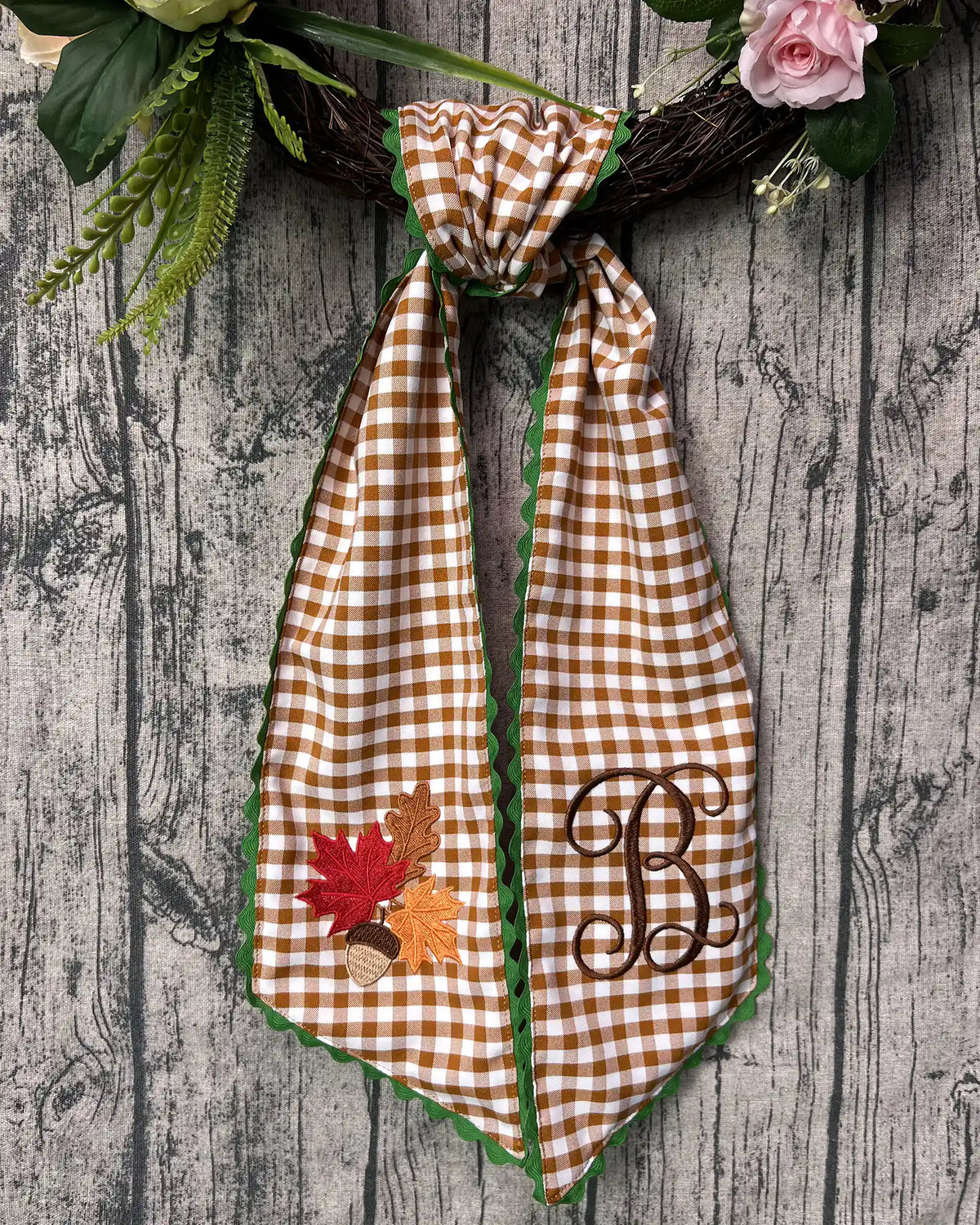 Brown gingham plaid wreath sash decorated on a floral wreath featuring embroidered autumn leaves, acorn, and monogram letter B.