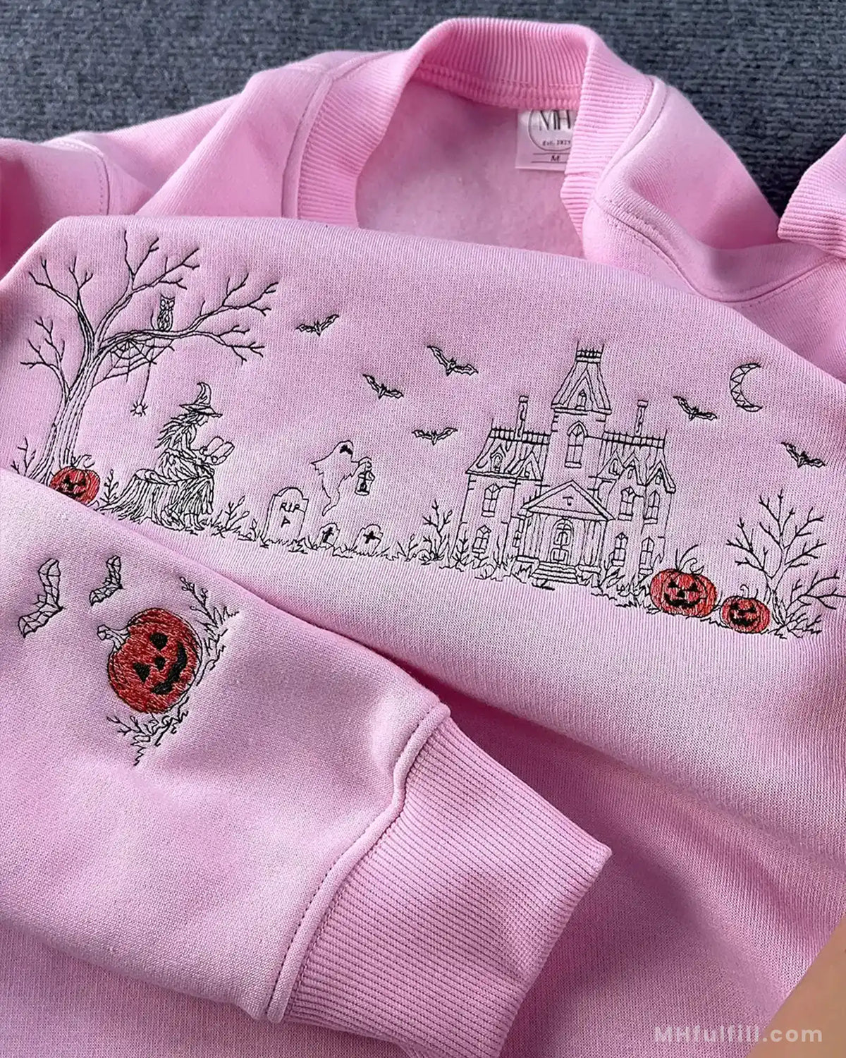 Detailed view of orange pumpkin and bat embroidery on the sleeve cuff of a pink Halloween sweatshirt.
