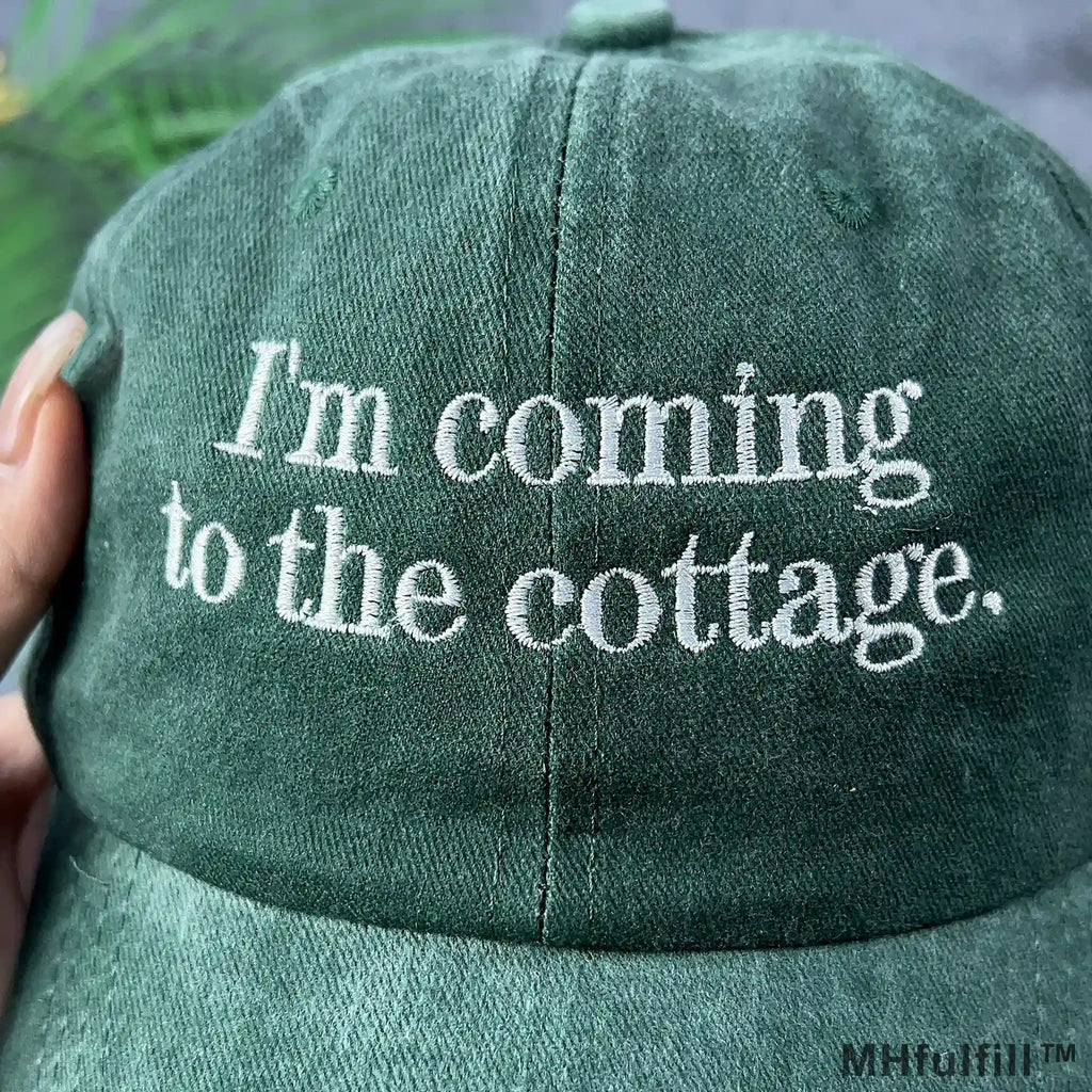 Close-up of white text embroidery reading I'm coming to the cottage on a vintage washed forest green dad hat.